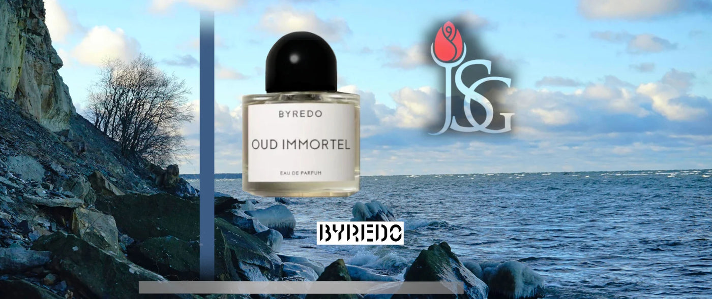 Image of an ocean side cliff with a semi-cloudy sky. In the foreground there is a bottle of Byredo's Oud Immortel perfume bottle with a vertical royal blue stripe and a horizontal while strip on the bottom. There is also the byredo logo and the jean smell good logo.
